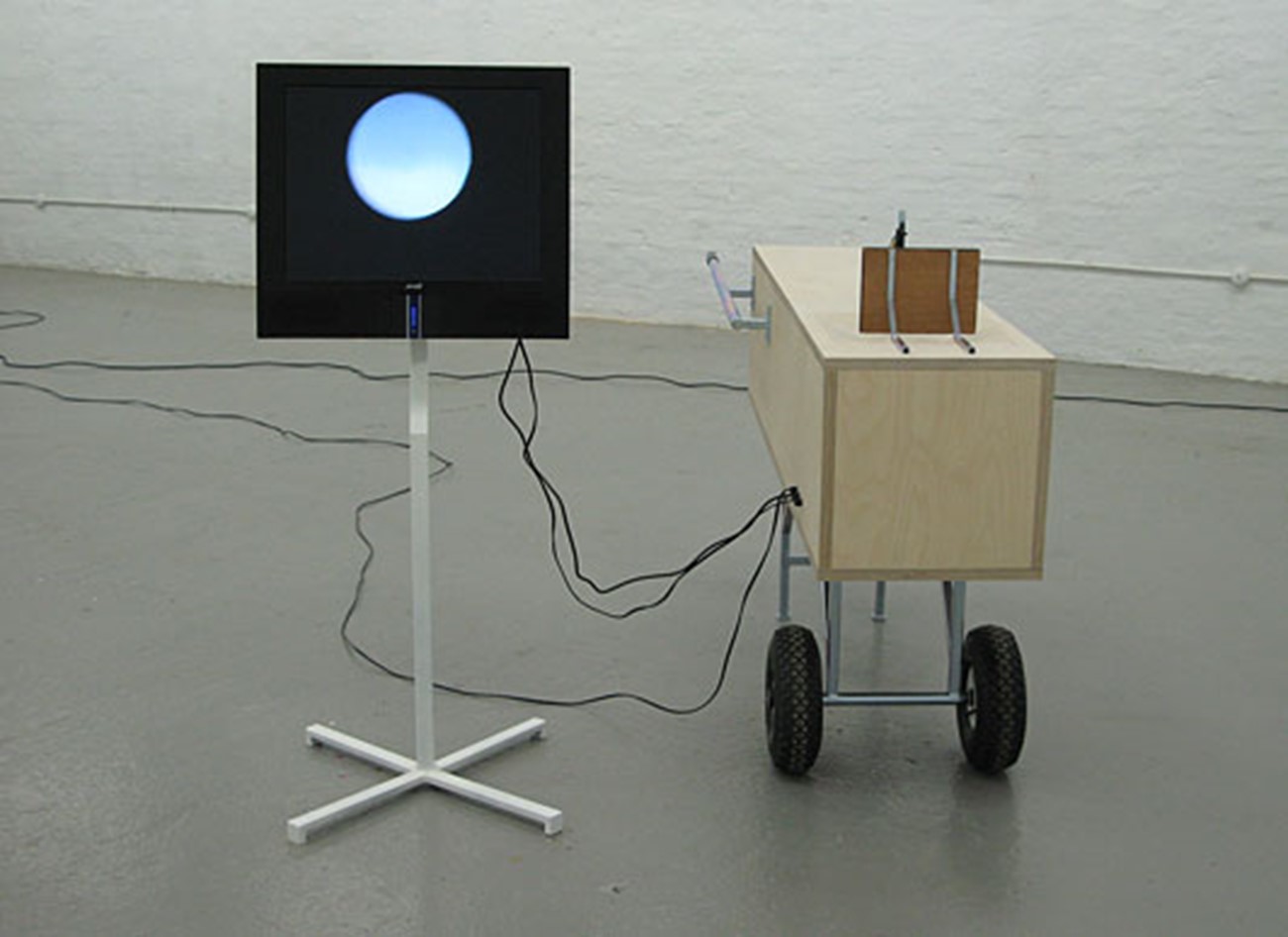 camera-based works — Måne uden navn (Moon with no name), 2008, Camera, iron, fan, screen , Variable dimensions (approx. 200 x 150 x 150 cm), A group of works by Henrik Menné is examinating the digital and analogue by underlining the transition from space to a twodimensional picture. A central element is the camera, pointing towards a set in movement. What happens in the three dimensions of the space is transmitted directly to the screen instead being stored digitally.