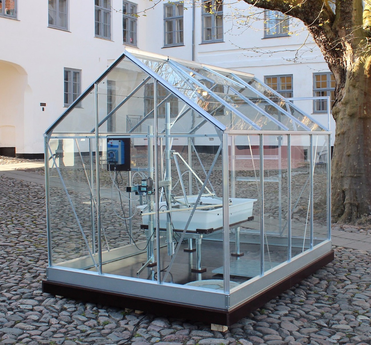 Afrundingsanordning (Installationview), 2015, Iron, glass, aluminum, plastic, wood and electronics , 207 x 193 x 257 cm, The machine drips a circle of citric acid on a grey green lime stone (an exclusive lime stone in high quality, 60x60x2). The acid gradually dissolves a trace in the stone's surface, which over time rounds off the initially squared stone. Viborg Kunsthal,.