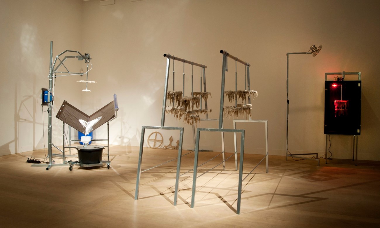 Fremstillingsenhed (Installation view), 2014, Build up around a machine, slowly producing white lumps of wood glue. On the floor is a beholder with wood glue, and on top of it a pump is placed, pumping the content out onto a tripod consisting of geometrical plates, mounted on a rotating metal stick. When the machine has being going on som time, the plates and the ironbars wil become covered with an organic lump of wood glue. Holstebro Kunstmuseum.