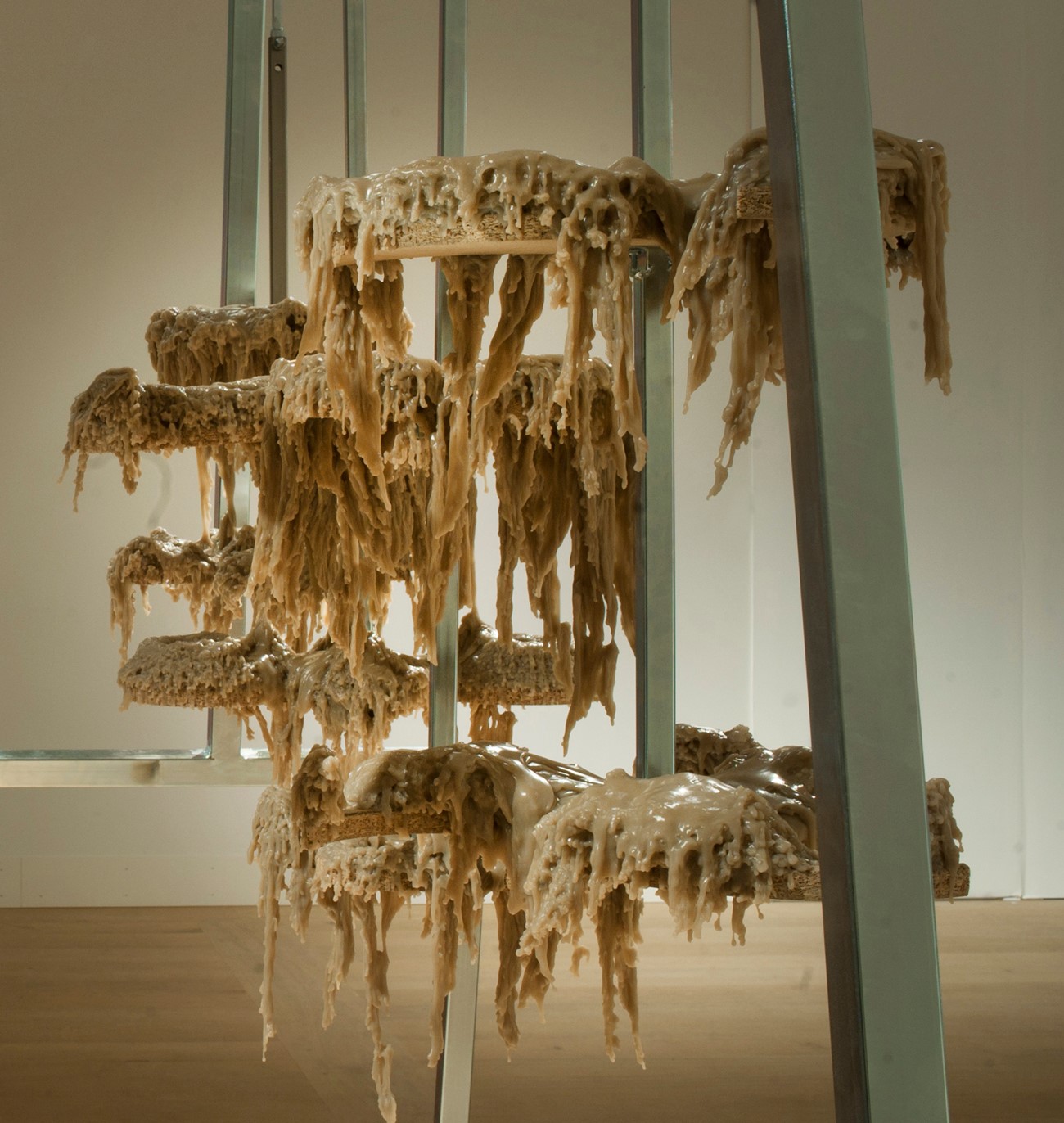 Fremstillingsenhed, 2014, Build up around a machine, slowly producing white lumps of wood glue. On the floor is a beholder with wood glue, and on top of it a pump is placed, pumping the content out onto a tripod consisting of geometrical plates, mounted on a rotating metal stick. When the machine has being going on som time, the plates and the ironbars wil become covered with an organic lump of wood glue. Holstebro Kunstmuseum.