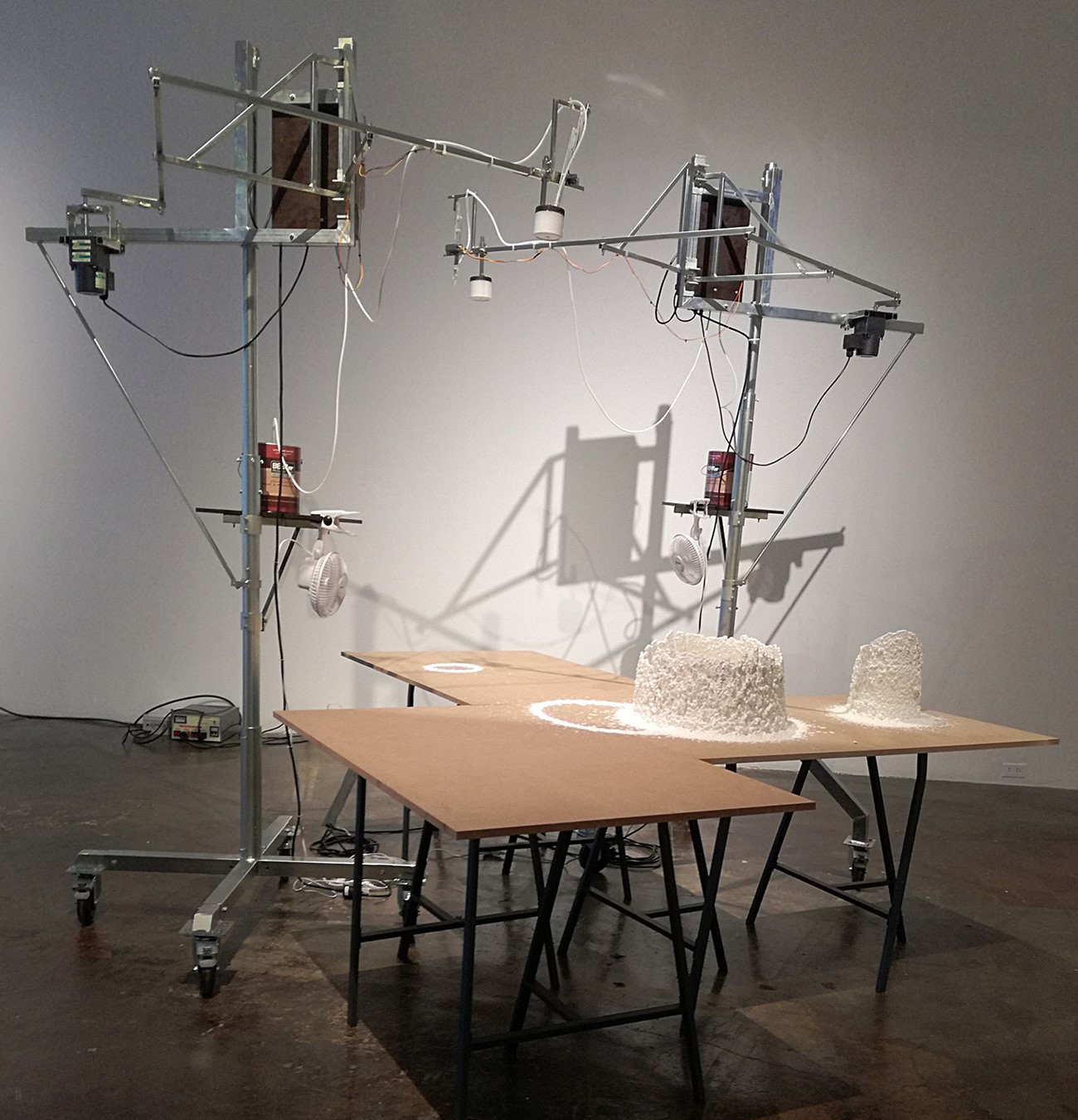 Paint Drip Device, 2018, Paint, ventilator, iron, heat element and electronics, 220 x 360 x 360 cm, The installation consists of two units which drip painting on a board. The device is constructed as an iron structure with a bucket of paint, a pump and a metal arm. The metal arm is fixed to a motor which makes it move around in a circle. The pump is connected to the paint bucket via a plastic hose. The outlet of the hose is fastened to the end of the metal arm. From there is very slowly drips painting on a table. The constant circular movements of the arm create a cylinder of dried painting.