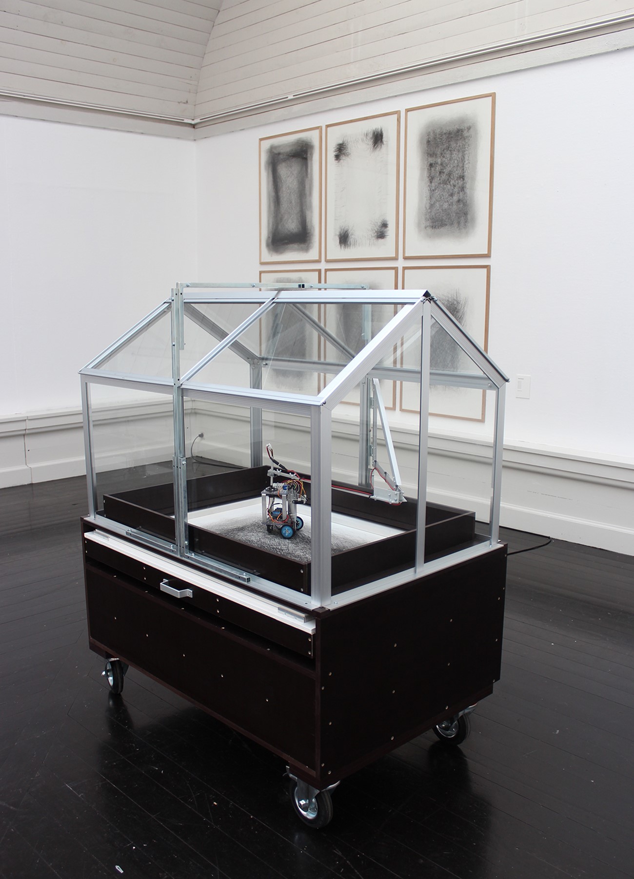 Tegningssystem TRK1, 2018, Iron, wood, paper and electronics, 160 x 80 x 120 cm, The drawing device consists of a wooden frame, a green house and a small robot with wheels which draws with a pen. The robot is controlled by a small computer which is installed on it. A couple of different sensors register where the robot is and how much is drawn on the paper under it. These inputs and the program decide how the drawing looks like. For every drawing a new program is uploaded to the computer.